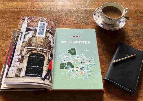 The book Doors of London sits open on a wooden table. The left page is a photograph of a black door with a soldier marching past. The right page is entitled Chapter 2: Westminster, with a map of Westminster on a mint green background. On the table beside the book is a cup of coffee in a blue-and-white china cup and saucer, and a black leather diary with a silver pen on top of it.