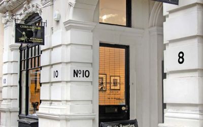 This colour photograph shows the Chris Beetles gallery on Ryder Street, St James’s, resplendent with rusticated façade, fluted pilasters and composite capitals. Large arched windows with black frames flank a grand entrance archway. Two black hanging signs with gold lettering identify the venue.