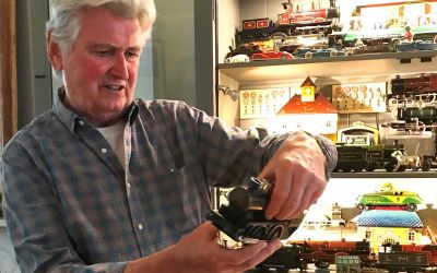 In this colour photograph, the designer Ian Logan holds a model steam locomotive with an illuminated cabinet behind him containing other models.