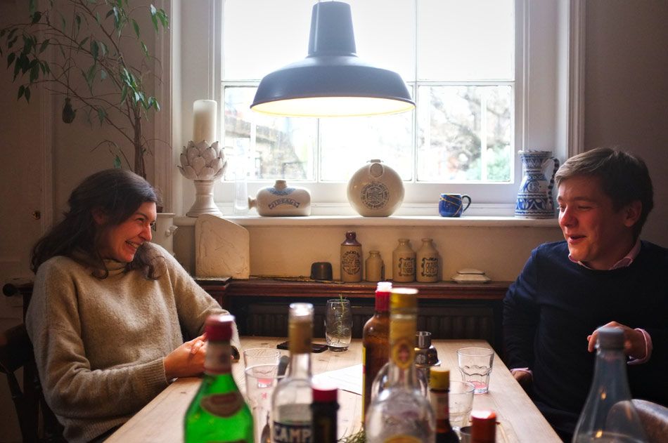 Two people chatting at a wooden kitchen table with multiple bottles of cocktail ingredients.