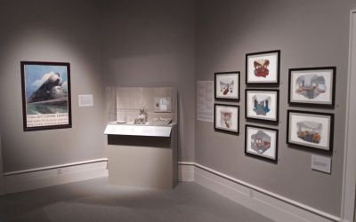 This colour photograph shows an exhibition room at the Albany Institute of History & Art featuring prints on the right-hand wall, a glass case containing silver dining-car cutlery centre and, on the far wall, a framed poster by Leslie Ragan depicting the streamlined 20th Century Limited locomotive.