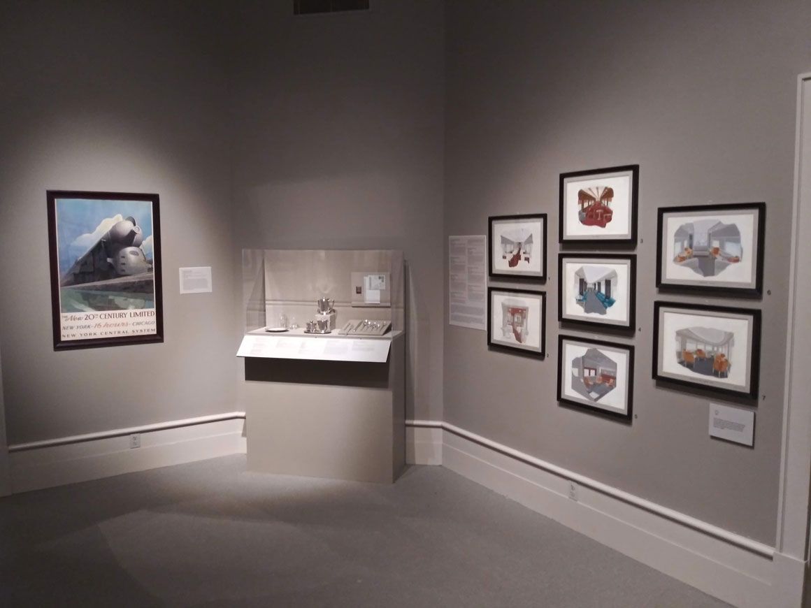 This colour photograph shows an exhibition room at the Albany Institute of History & Art featuring prints on the right-hand wall, a glass case containing silver dining-car cutlery centre and, on the far wall, a framed poster by Leslie Ragan depicting the streamlined 20th Century Limited locomotive.