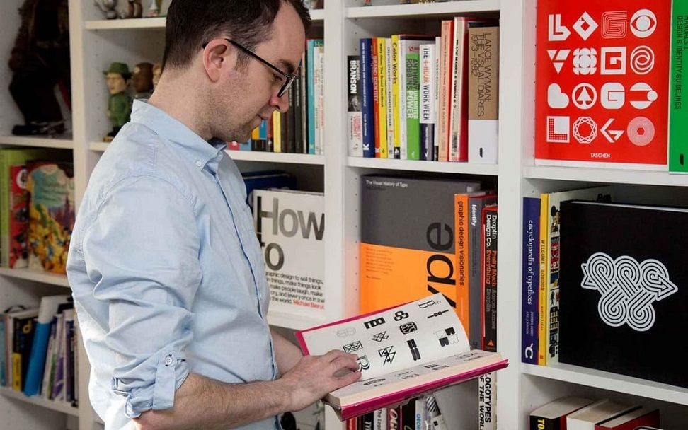 The logo designer Ian Paget, wearing a blue shirt, prepares to geek out with logos as he peruses one of his many books on logo design for inspiration.