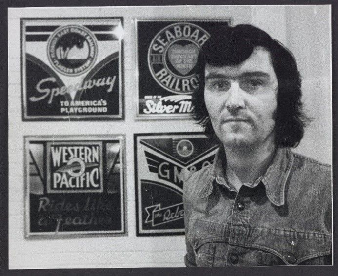 Black and white photograph of Ian Logan standing beside four American railroad logos he designed in the 1970s.