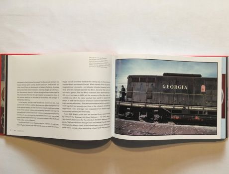 In this colour photograph taken by Ian Logan in the 1980s a GP 7 diesel locomotive of the Georgia Railroad inches forward with a lookout standing on the forward platform. Painted dark grey, it carries the name Georgia painted in white serif capitals and the badge of the Georgia Railroad with three white pillars on a red background labelled Courtesy, Safety and Service supporting a lintel and arch emblazoned with the name Georgia RR, Old Reliable. 
