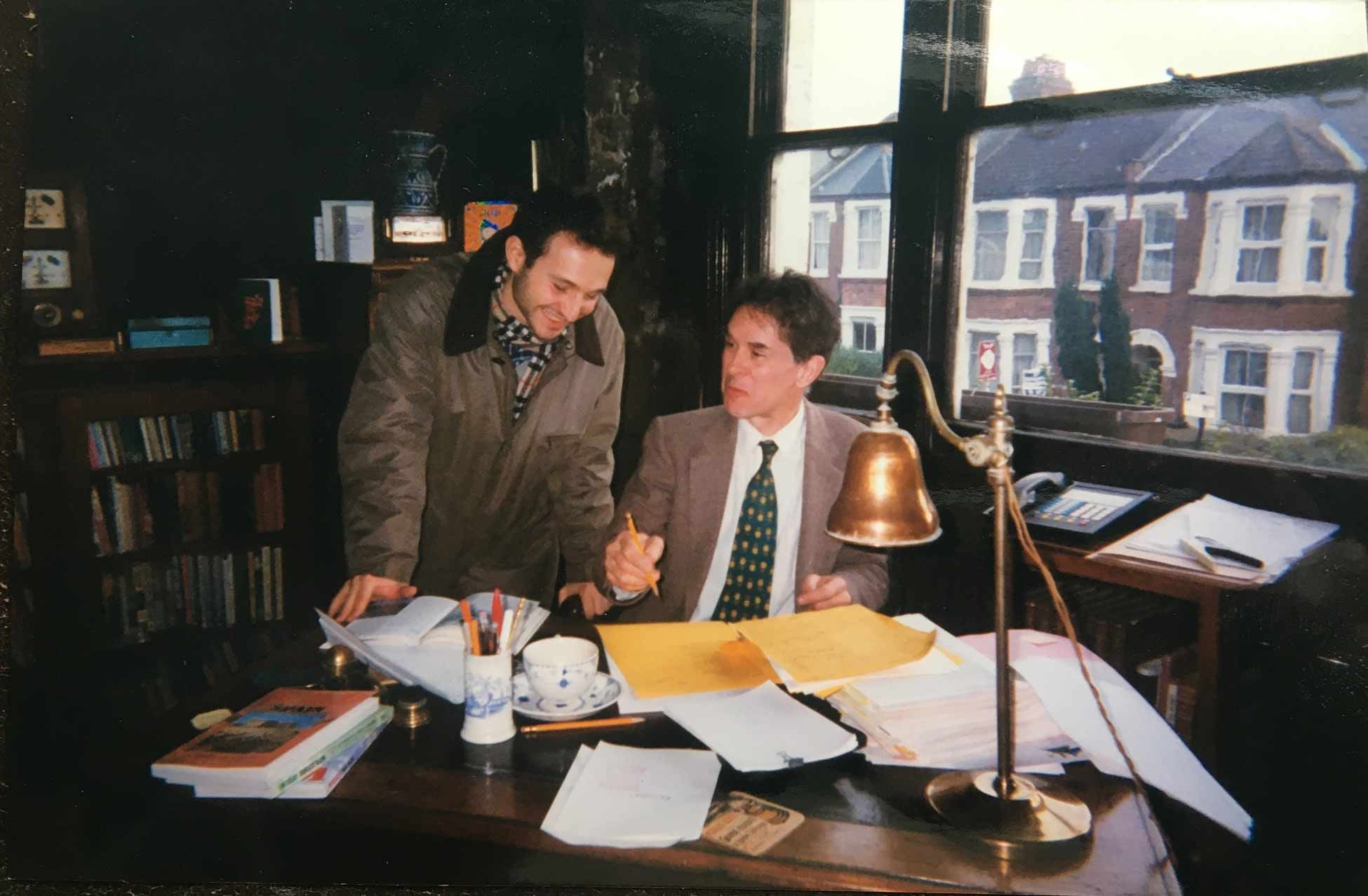 In a dark chocolate brown study, the Publisher sits at his pedestal desk, pencil in hand, and discusses manuscripts with a member of staff.