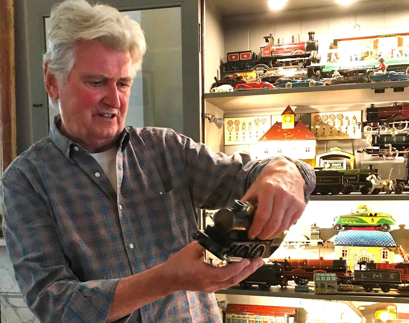 In this colour photograph, the designer Ian Logan holds a model steam locomotive with an illuminated cabinet behind him containing other models.