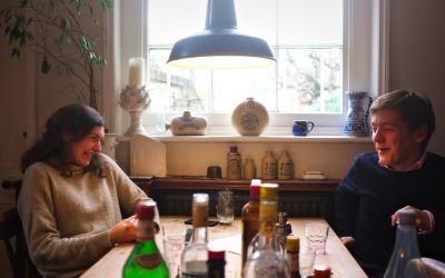 Two people chatting at a wooden kitchen table with multiple bottles of cocktail ingredients.