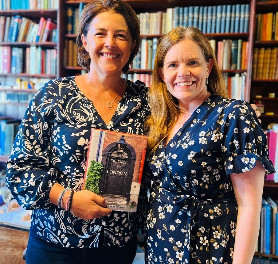 The door detectives Cath Harries and Melanie Backe-Hansen, dressed in similar blue-and-white patterned tops, hold a mock-up of their book Doors of London with an 18th-century door in Spitalfields on the cover.