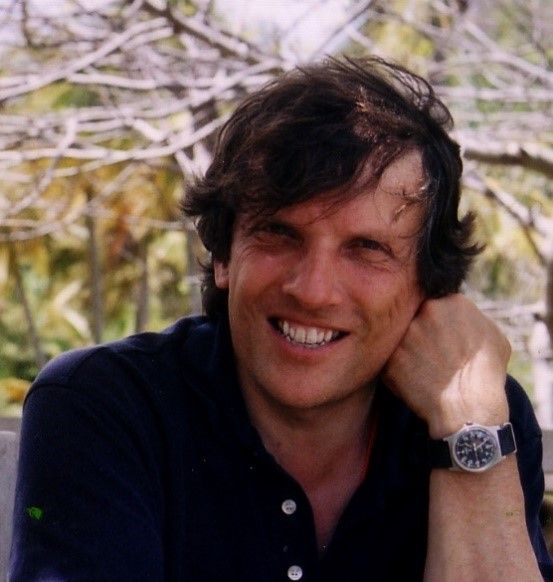 In this colour photograph the design critic Jonathan Glancey, co-author of Logomotive: Railroad Graphics and the American Dream with Ian Logan and Norman Foster, smiles at the camera as he sits on a garden bench with a backdrop of trees.