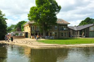A colour photograph of the Heath Robinson Museum and West House is taken from the far side of the adjacent lake. Visitors sit at café tables outdoors and a family feeds ducks at the water’s edge.