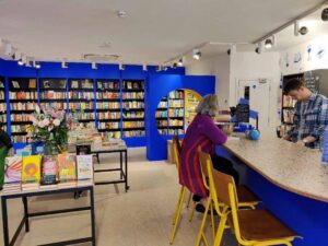 A staff member stands behind the big blue counter of Backstory while a customer enjoys a pot of tea with her new book.