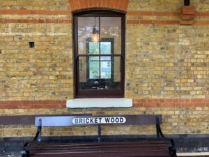 In the centre of this photograph taken on the platform at Bricket Wood Station is a sash window painted black and beneath it a bespoke wooden bench with the letters Bricket Wood set into the back rest.