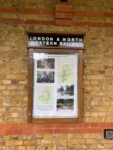 On the platform wall at Bricket Wood Station a glass-fronted poster cabinet with a brown frame is headed London & North Western Railway in white capitals on a black background.