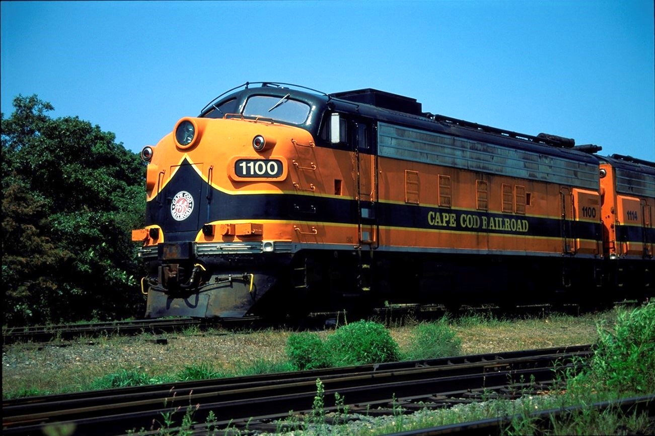 An F-Type Loco painted in orange, black and yellow livery hauls an excursion train on the Cape Cod Central Railroad.