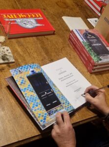 A book lies open on a wooden table. The page to the left features a black door surrounded by walls with a bright blue-and-yellow pattern. The page to the right is the title page. It reads ‘Doors of London, Styles, Stories, Art and Architecture, Cath Harries and Melanie Backe-Hansen’. Using a black pen, a hand signs ‘Cath Harries’ on the title page. Three books entitled ‘Doors of London’ are stacked to the right of the main book.