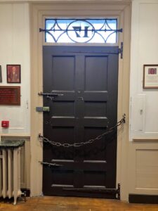 In this colour photograph of the back of the door at Dr Johnson’s house, you can see the spiked iron bar behind the fanlight, enormous T hinges, and a heavy chain secured on a corkscrew latch.