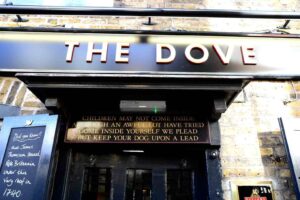 Pictured is the entrance of The Dove pub. Above the door are two signs. The main sign features the name of the pub in capitalized white font. The sign below reads: Children may not come inside although an awful lot have tried. Come inside yourself we plead, but keep your dog upon a lead. This is written in gold capitalized lettering against a black background. To the left of the door is a chalkboard with chalk writing on it, reading: Did you know that James Thompson penned Rule Britannia under this very roof in 1740?