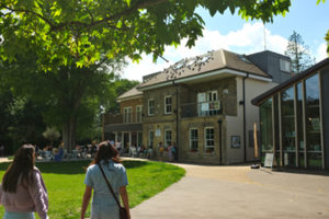 A colour photograph of the Heath Robinson Museum in the Pinner Memorial Park is taken from beneath the canopy of a nearby tree. The Museum flanks West House, where a busy café has spilled into the shade outside.