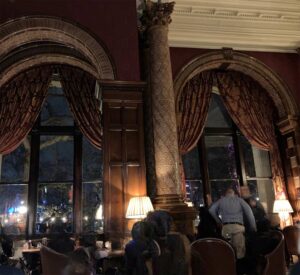 In a colour photograph shot in the bar at the National Liberal Club, swagged curtains and dark wood panelling are warmly lit by table lamps as members chat in comfort at spaciously laid out tables.