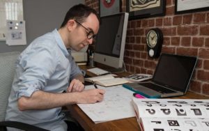 In this colour photograph the logo designer Ian Paget, wearing a blue shirt with the sleeves rolled up, gets to work on sketching new logo ideas.