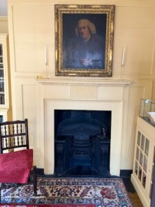 In this colour photograph in the library, an oil portrait of Dr Johnson in a gilt frame hangs over the fireplace and a comfortable rush seat chair with a red cushion stands ready for a reader.