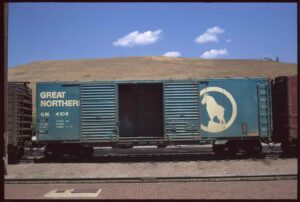 In this photograph by Ian Logan, a goods van stands on tracks in front of a large hill of parched grass. On the left of the van’s open doors is the name of the Great Northern Railway painted in white, whilst on the right is the railway’s logo, a mountain goat atop a rocky crag.