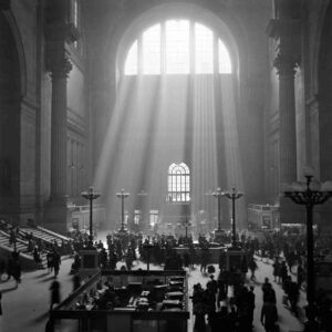 This black-and-white photograph depicts Pennsylvania station with its huge window, casting rays of light into the interior. Inside are vintage lamps and commuters making their way through the station.