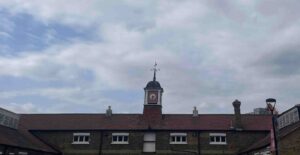 Photographed is the roof of the historic Ram Brewery stables. Constructed of red tiles and sloping down over a London stock brick building, a clock tower rises from the roof’s centre, with a bell roof and a spire leading up to a weathervane. Grey clouds are coming in from the right, covering the light blue sky visible on the left.
