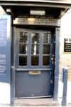 Pictured is a wide grey blue door with multiple small square windows and a brass letterbox. The sign above the door reads: Children may not come inside although an awful lot have tried. Come inside yourself we plead, but keep your dog upon a lead.