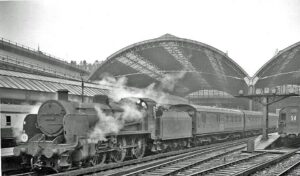 This black-and-white photograph depicts a steam train exiting Victoria Station, followed by wisps of smoke.