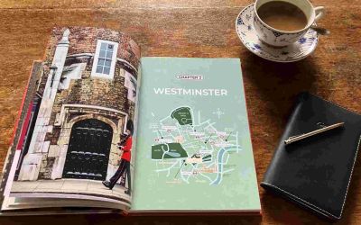 The book Doors of London sits open on a wooden table. The left page is a photograph of a black door with a soldier marching past. The right page is entitled Chapter 2: Westminster, with a map of Westminster on a mint green background. On the table beside the book is a cup of coffee in a blue-and-white china cup and saucer, and a black leather diary with a silver pen on top of it.