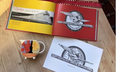 In this colour photograph, a copy of the book ‘Logomotive’ lies open at a page with a deep red background carrying the logo of the ‘Zephyr’ express train while alongside is a limited-edition black-and-white print of the same logo and an Art Deco milk jug.