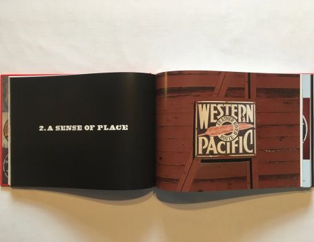 In this colour photograph taken by the railfan designer Ian Logan, a rusting enamel sign screwed to the side of a wooden boxcar carries the name Western Pacific picked out in white serif capitals on a black background with a red feather pointing through a central circle which carries the legend Feather River Route. 