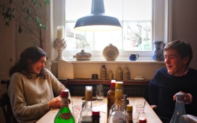 Two people chatting at a wooden kitchen table with multiple bottles of cocktail ingredients.
