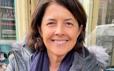 In this colour photograph Cath Harries looks at the camera and smiles as she pauses for a photograph while out door hunting, wearing a dark blue leather jacket and a scarf.